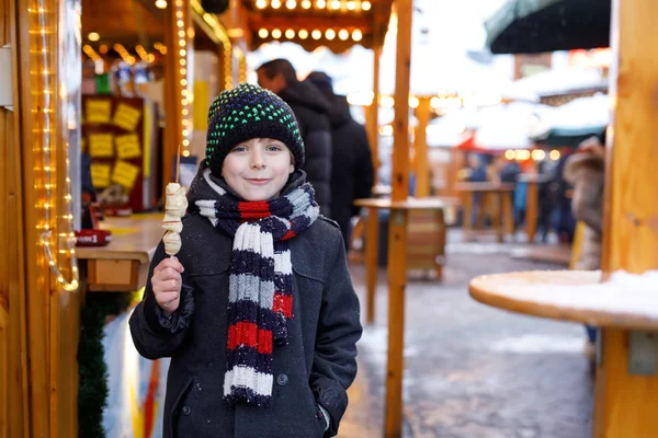 Petit garçon mignon mangeant des fruits couverts de chocolat blanc sur brochette sur le marché de Noël allemand traditionnel. Enfant heureux sur le marché familial traditionnel en Allemagne, Munich pendant la journée enneigée . — Photo