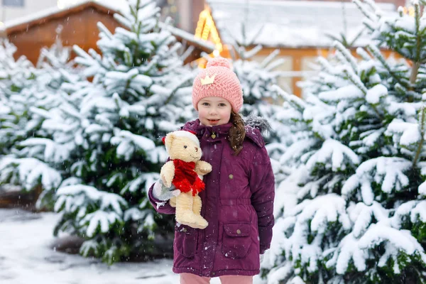 Linda niña sonriente en el mercado de árboles de Navidad. Niño feliz en ropa de invierno y juguete eligiendo el árbol de Navidad en el mercado de Navidad con luces en el fondo en el día de nieve de invierno . — Foto de Stock