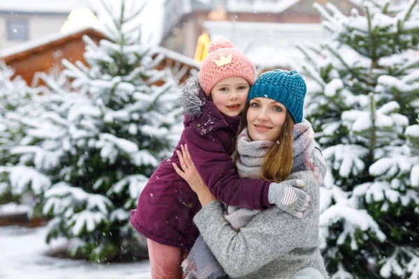Jolie petite fille souriante et mère sur le marché des arbres de Noël. Joyeux enfant, fille et jeune femme en vêtements d'hiver choisissant arbre de Noël sur le marché de Noël avec des lumières sur le fond le jour de neige d'hiver — Photo