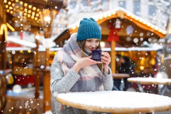 Beautiful young woman drinking hot punch, mulled wine on German Christmas market. Happy girl in winter clothes with lights on background on winter snow day in Munich, Germany.