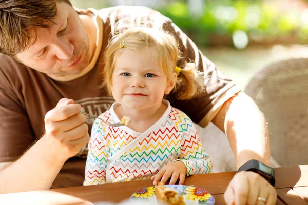 Young middle-aged father feeding cute little toddler girl in restaurant. Adorable baby child learning eating from spoon. Happy healthy family in an outdoor cafe in summer time, eating cake — Stock Photo, Image