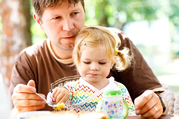 Young middle-aged father feeding cute little toddler girl in restaurant. Adorable baby child learning eating from spoon. Happy healthy family in an outdoor cafe in summer time, eating cake — Stock Photo, Image