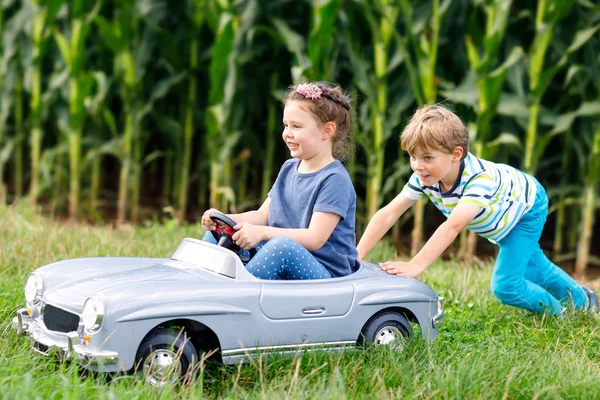 Двое счастливых детей, играющих с большой старой игрушечной машиной в летнем саду, на улице. Мальчик водит машину с маленькой девочкой внутри. Смеющиеся и улыбающиеся дети. Семья, детство, образ жизни.. — стоковое фото
