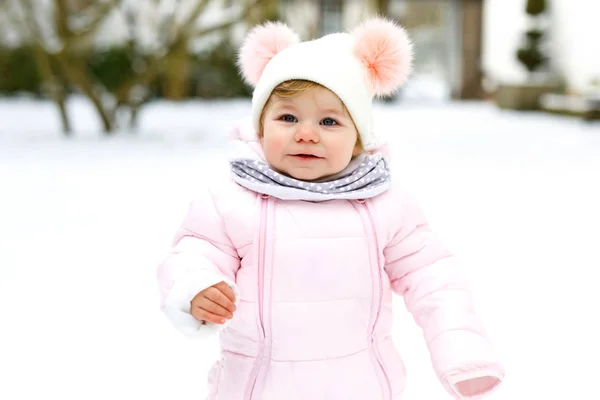 Adorável menina fazendo primeiros passos ao ar livre no inverno. Criança bonito aprender a andar. Criança se divertindo no dia de neve fria. Vestindo roupas quentes rosa bebê e chapéu com bobbles. — Fotografia de Stock