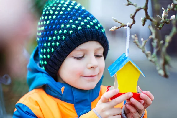 Little kid boy feeding birds in winter. Child hanging colorful selfmade bird house on tree on frosty cold day. Happy preschooler in colorful wam clothes. Selective focus on child and feeder with seeds — Stock Photo, Image