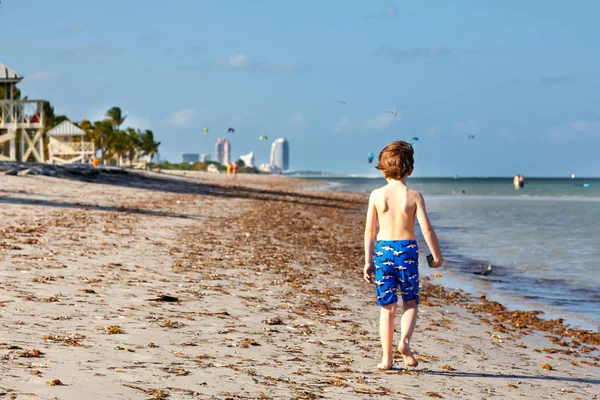Niño rubio divirtiéndose en la playa de Miami, Key Biscayne. Feliz niño lindo saludable jugando con arena y corriendo cerca del océano. Palmeras, casa de seguridad y arena blanca —  Fotos de Stock