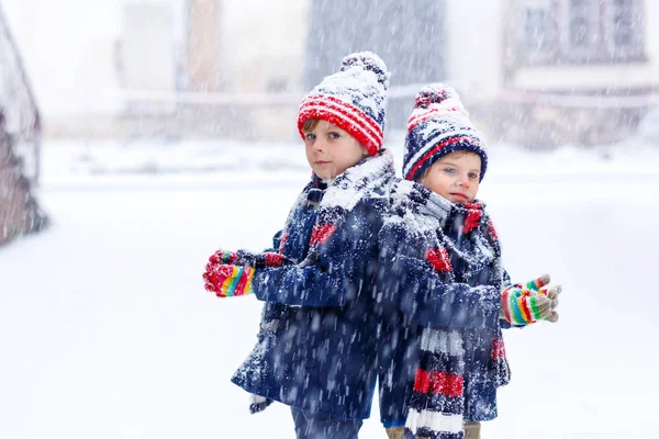 Två små pojkar i färgglada modekläder som leker utomhus under starkt snöfall. Aktiv fritid med barn på vintern under kalla dagar. Glada syskon och tvillingar har kul med första snön. — Stockfoto