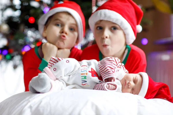 Een week oud pasgeboren babymeisje en twee broers en zussen kid jongens in Santa Claus hoeden in de buurt van de kerstboom met kleurrijke garland lichten op achtergrond. Close-up van drie kinderen, het vieren van Kerstmis en gelukkige familie. Stockfoto