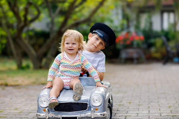 Dvě šťastné děti si hrají s velkým starým autíčkem v letní zahradě, venku. Chlapec řídí auto s malou batolatou holčičkou uvnitř. Smějící se a usměvavé děti. Rodina, dětství, životní styl — Stock fotografie