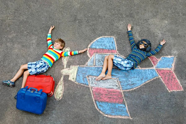 Two little children, kids boys having fun with with airplane picture drawing with colorful chalks on asphalt. Friends painting with chalk and going on vacations dreaming of pilot profession.