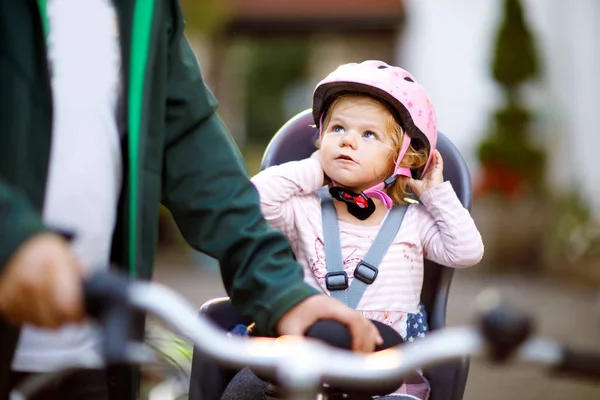 Portrait of little toddler girl with security helmet on the head sitting in bike seat and her father or mother with bicycle. Safe and child protection concept. Family and weekend activity trip.