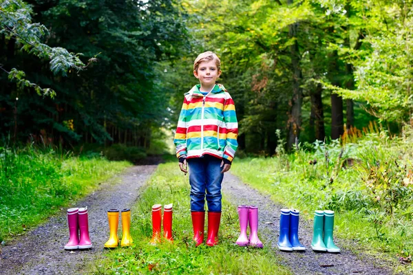 Liten unge pojke och grupp av färgglada regn stövlar. Blont barn står i höst skog. Närbild av schoolkid och olika gummistövlar. Skor och mode för regniga hösten — Stockfoto