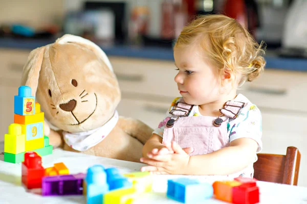 Gadis balita manis dengan kelinci mewah favorit bermain dengan mainan pendidikan di kamar bayi. Anak yang bahagia dan sehat bersenang-senang dengan balok plastik yang berbeda warna-warni di rumah. Lucu bayi belajar menciptakan. — Stok Foto