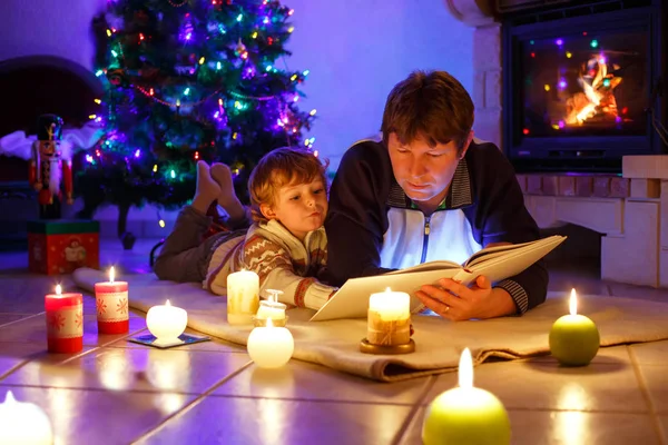 Father and cute little toddler boy reading book by chimney, candles and fireplace. — Stock Photo, Image