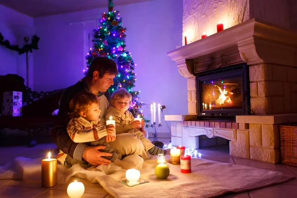 Ojciec i dwóch małych chłopców siedzących przy kominku, świecach i kominku i patrzących w ogień. — Zdjęcie stockowe