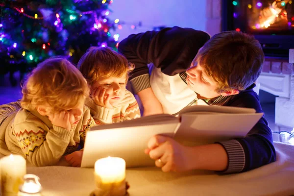 Father and two little toddler boys reading book by chimney, candles and fireplace. — Stock Photo, Image