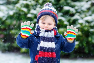 Renkli kış kıyafetleri içinde, kar yağarken dışarıda oynayıp eğlenen sevimli, komik bir çocuk.