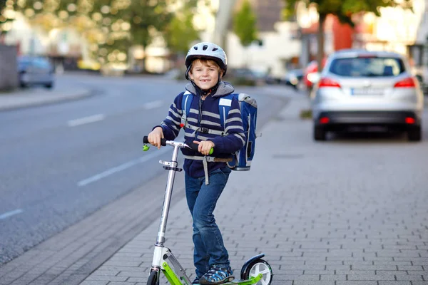 Bambino della scuola attiva in casco di sicurezza cavalcando con il suo scooter in città con lo zaino nella giornata di sole. Bambino felice in vestiti colorati in bicicletta sulla strada per la scuola. — Foto Stock