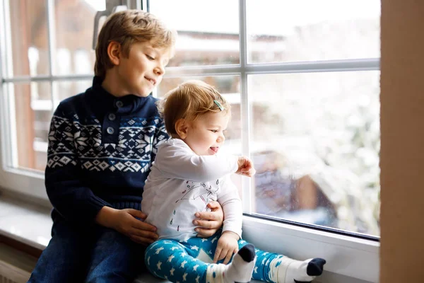 Happy adorable kid boy and cute baby girl sitting near window and looking outside on snow on Christmas day or morning — Stock Photo, Image
