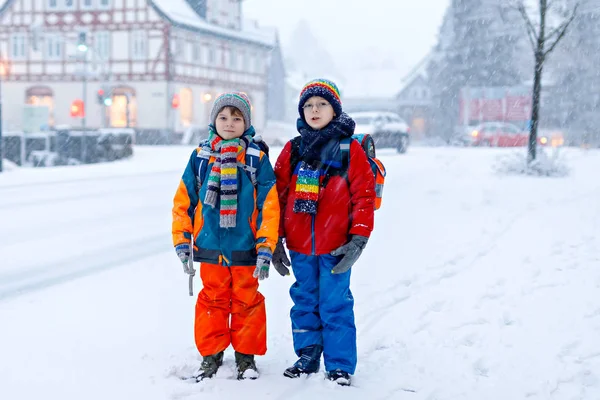 Dva malí kluci ze základní třídy chodili během sněžení do školy. Šťastné děti se baví a hrají si s prvním sněhem. Sourozenci přátelé s batohem v barevných zimních šatech. — Stock fotografie