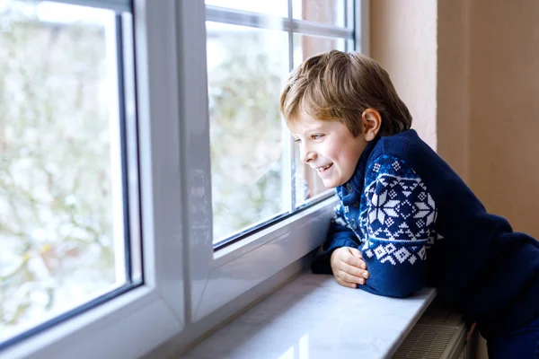 Menino criança adorável feliz sentado perto da janela e olhando para fora na neve no dia de Natal ou de manhã . — Fotografia de Stock