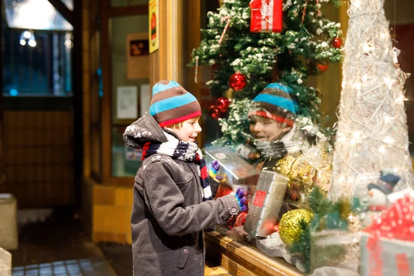 Funny happy child in fashion winter clothes making window shopping decorated with gifts, xmas tree — Stock Photo, Image