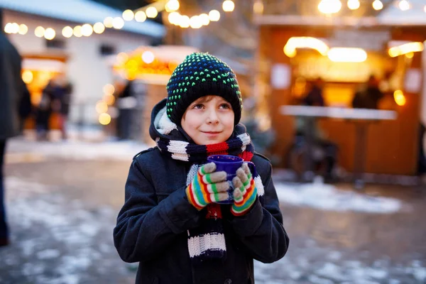 Niño lindo bebiendo ponche de niños calientes o chocolate en el mercado de Navidad alemán. Niño feliz en el mercado familiar tradicional en Alemania, niño risueño en ropa de invierno colorida — Foto de Stock