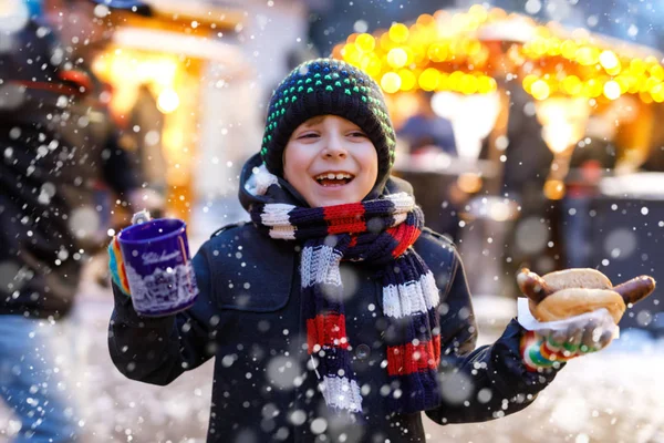Little cute kid boy eating German sausage and drinking hot children punch on Christmas market — Stock Photo, Image