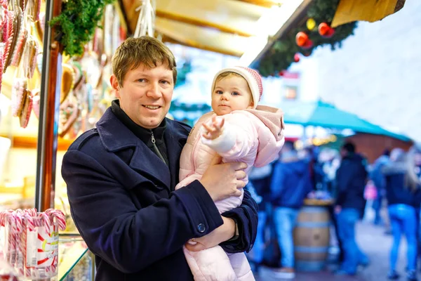 Père d'âge moyen tenant bébé fille près de stand doux avec du pain d'épice et des noix. Bonne famille sur le marché de Noël en Allemagne. Jolie fille manger un cookie appelé Lebkuchen. Célébration Noël vacances. — Photo