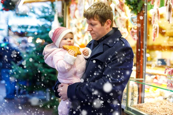 Отец средних лет держит дочку рядом со сладким ларьком с пряниками и орехами. Счастливая семья на рождественском рынке в Германии. Милая девушка ест печенье по имени Лебкучен. Празднование Рождества Христова. — стоковое фото