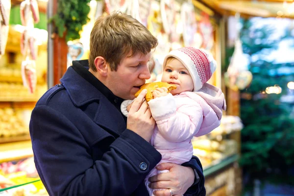 Pai de meia idade segurando bebê filha perto de carrinho doce com pão de gengibre e nozes. Família feliz no mercado de Natal na Alemanha. Menina bonito comer biscoito chamado Lebkuchen. Celebração Natal feriado. — Fotografia de Stock