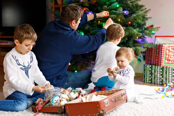 Zwei kleine Jungen und ein entzückendes Mädchen schmücken den Weihnachtsbaum mit altem Spielzeug und Bällen. Papa im Hintergrund — Stockfoto