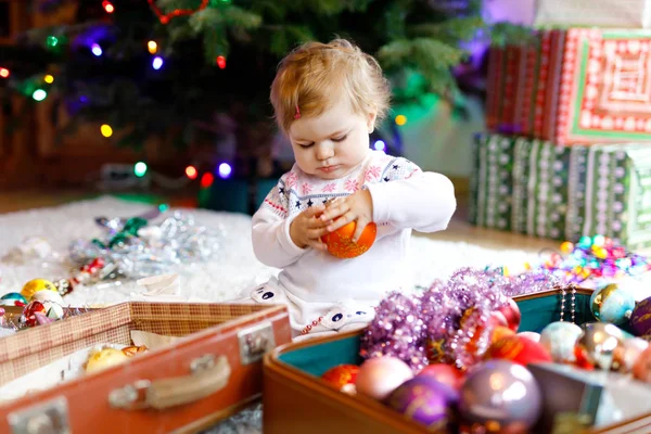 Очаровательная девочка держит красочные старинные рождественские игрушки и мяч в милых руках. Маленький ребенок в праздничной одежде украшая елку — стоковое фото