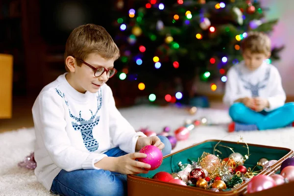 Beautiful kid boy with eye glasses and colorful vintage xmas toys and balls in old suitcase. Little child, school boy in festive clothes decorating Christmas tree. Brother on background