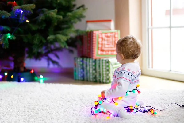 Adorable baby girl holding colorful lights garland in cute hands. Little child in festive clothes decorating Christmas tree — Stock Photo, Image