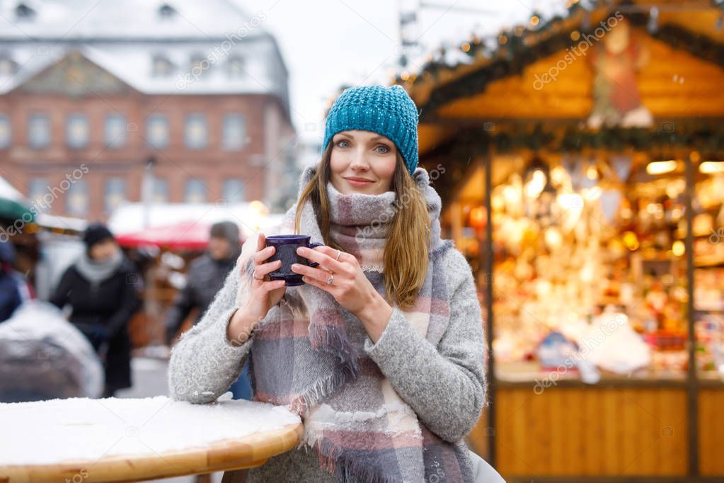 Beautiful young woman drinking hot punch, mulled wine on German Christmas market. Happy girl in winter clothes with lights on background on winter snow day in Hanau, Germany