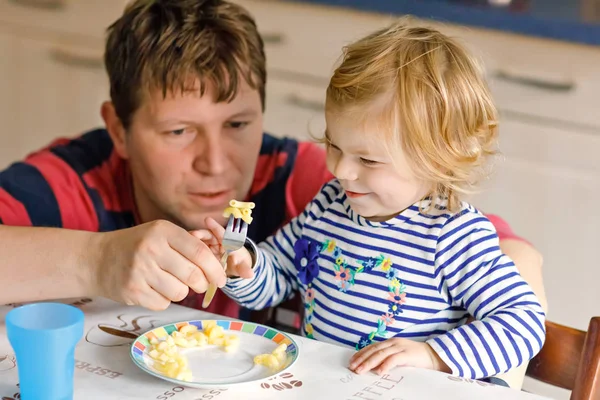 Schattig baby meisje eten noodle, pasta macaroni. Vader helpen dochter leren lepel gebruiken. Schattig gezonde peuter kind zit in de kinderstoel, leren om te eten door zelf met papa in de binnenlandse keuken — Stockfoto