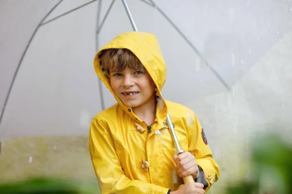 Bellissimo bambino sulla strada per la scuola a piedi durante sleet, pioggia forte e neve con un ombrello nella giornata fredda. Bambino felice e gioioso in abiti casual colorati cappotto giallo moda — Foto Stock