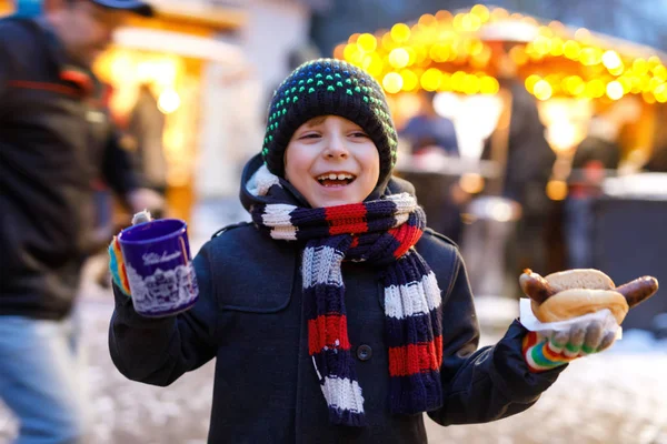 Little cute kid boy eating German sausage and drinking hot children punch on Christmas market — Stock Photo, Image