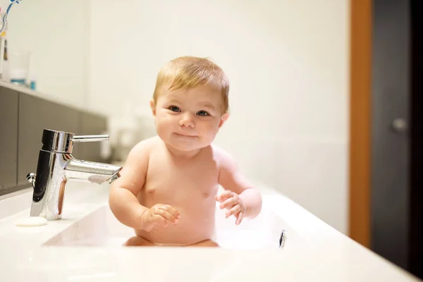 Schattige schattig baby nemen bad in wassen gootsteen en waterkraan te grijpen. Gezond meisje met grote blauwe ogen hebben — Stockfoto