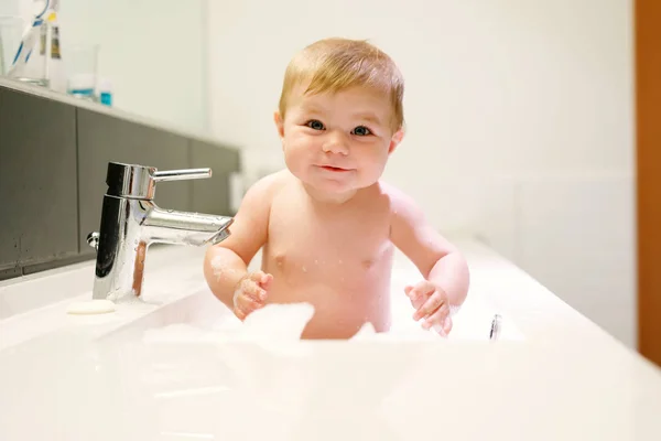 Roztomilé roztomilé dítě se koupe v umyvadle a hraje si s vodou a pěnou — Stock fotografie