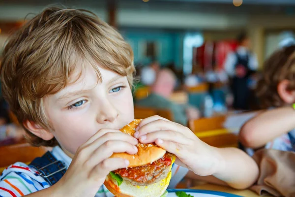 Ładny chłopak przedszkola zjada hamburger siedzi w stołówce szkolnej — Zdjęcie stockowe