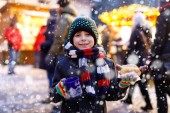 Der kleine süße Junge isst Bratwurst und trinkt auf dem Weihnachtsmarkt heißen Kinderpunsch. Glückskind auf dem traditionellen Familienmarkt in Deutschland, München. Lachender Junge in bunter Winterkleidung