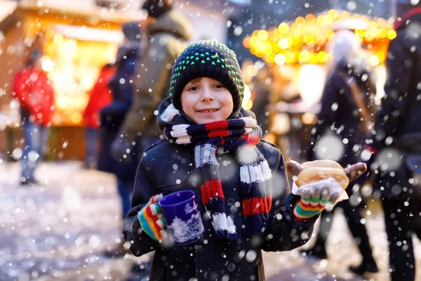 Der kleine süße Junge isst Bratwurst und trinkt auf dem Weihnachtsmarkt heißen Kinderpunsch. Glückskind auf dem traditionellen Familienmarkt in Deutschland, München. Lachender Junge in bunter Winterkleidung — Stockfoto