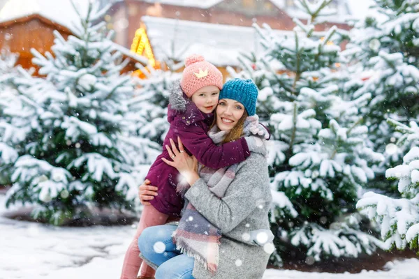 Cute little smiling kid girl and mother on christmas tree market. Happy child, daughter and young woman in winter clothes choosing xmas tree on xmas market with lights on background on winter snow day — Stock Photo, Image