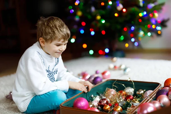 Vacker pojke och färgglada vintage xmas leksaker och bollar i gamla resväska. Ett litet barn, skolpojke i festliga kläder som dekorerar julgranen. Ungen ler mot kameran. — Stockfoto