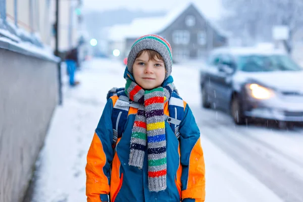 Kisiskolás fiú, általános osztályból, aki hóesés idején sétál az iskolába. Kora reggel és havas utcák a városban. Diák hátizsák vagy táska színes téli ruhák. — Stock Fotó