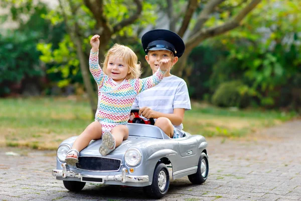 Dvě šťastné děti si hrají s velkým starým autíčkem v letní zahradě, venku. Chlapec řídí auto s malou batolatou holčičkou uvnitř. Smějící se a usměvavé děti. Rodina, dětství, životní styl — Stock fotografie