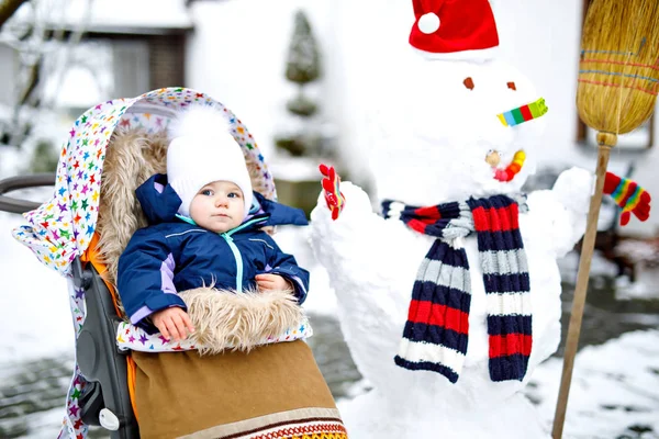 Cute little beautiful baby girl siedzi w wózku lub wózek w zimowy dzień ze śniegiem i bałwanem. Szczęśliwy uśmiech dziecka w ciepłych ubraniach, moda stylowe dziecko.. — Zdjęcie stockowe