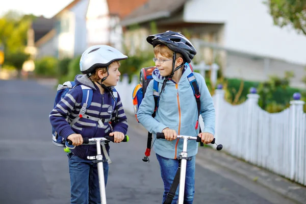 Dwóch chłopców w kasku jeździ z hulajnogą w mieście z plecakiem w słoneczny dzień. Szczęśliwe dzieci w kolorowe ubrania na rowerze w drodze do szkoły. — Zdjęcie stockowe